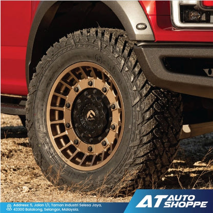 Close-up of a red vehicle with a large off-road tire and bronze rim on a dirt road, featuring the AT Auto Shoppe logo.