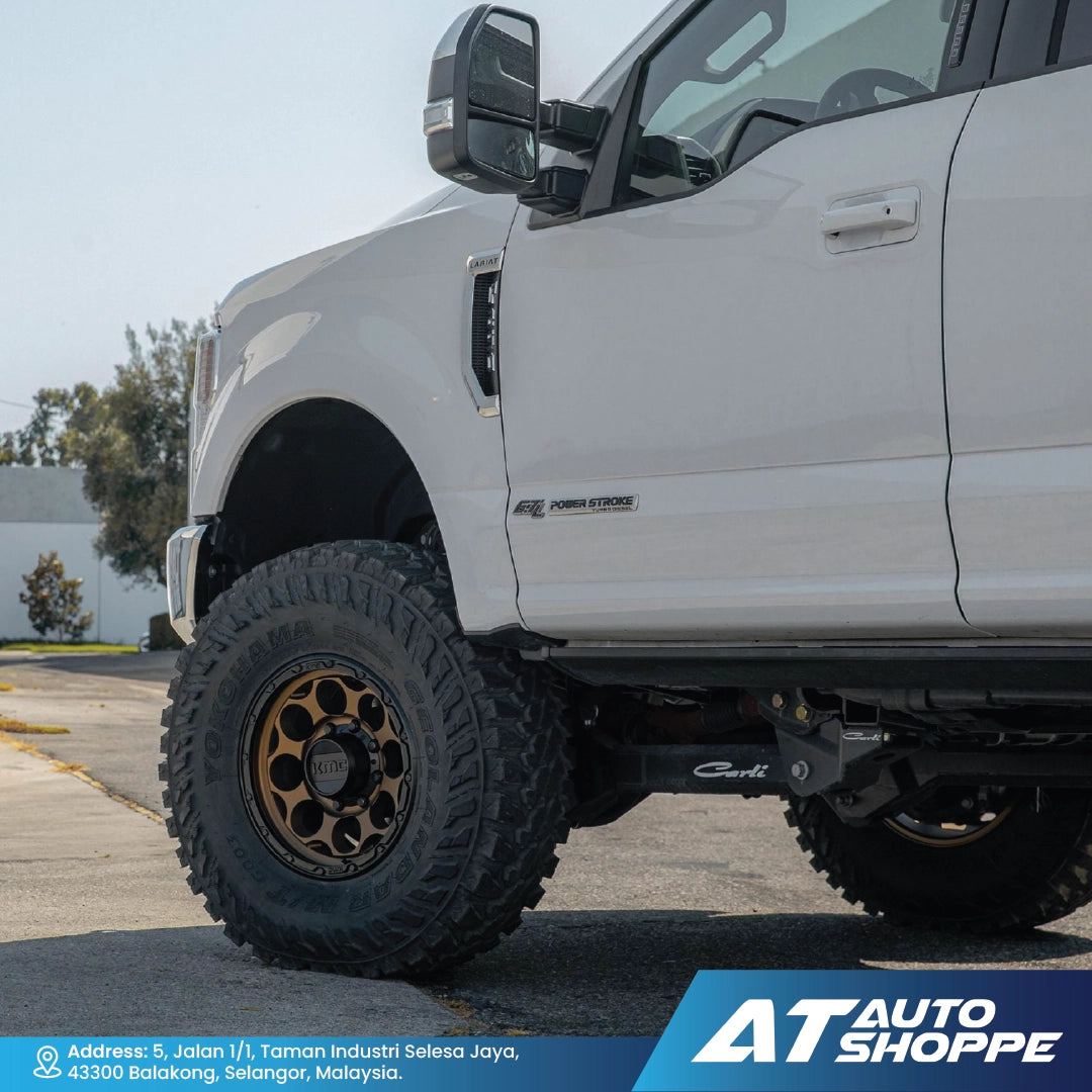 White pickup truck with large off-road tires on a paved surface, featuring AT Auto Shoppe branding.