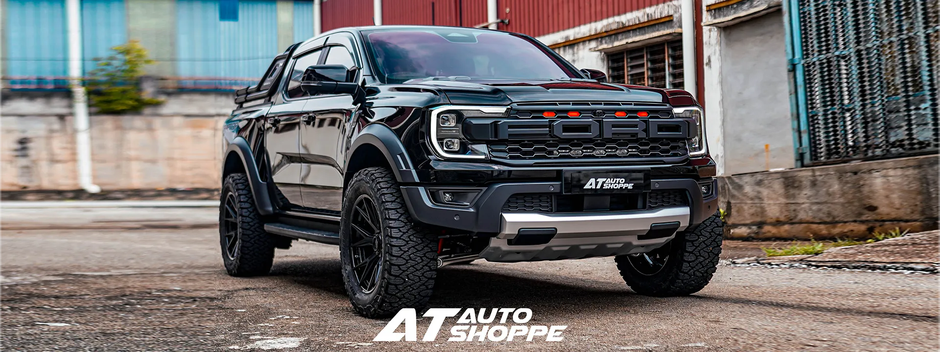 A black Ford Ranger pickup truck with off-road modifications, including a large front grille, heavy-duty front bumper, and large off-road tires, parked on a paved street in front of industrial buildings.