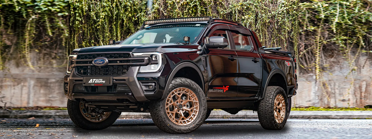 A black Ford Ranger pickup truck with a custom front bumper, roof light bar, and large bronze wheels, parked outdoors in front of a stone wall covered in green ivy.