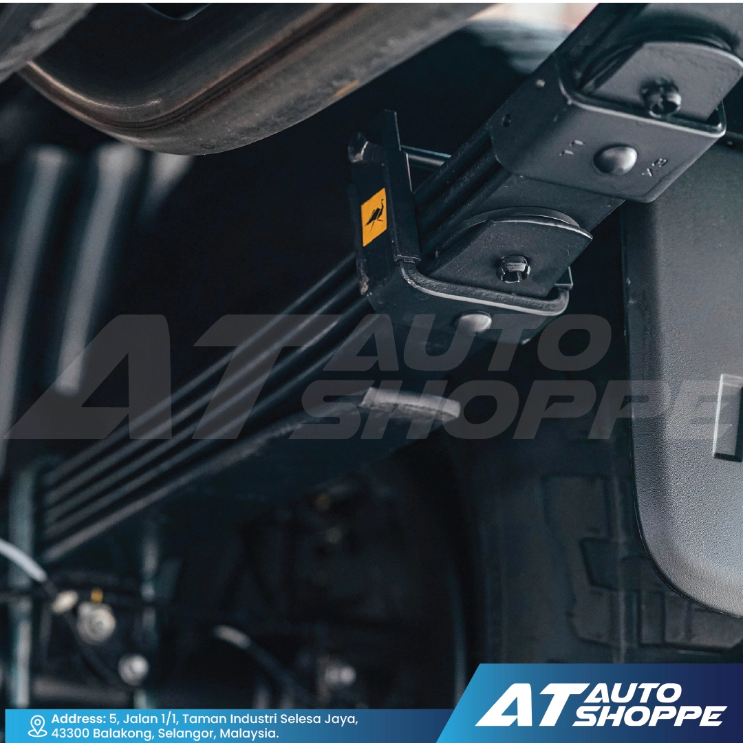 Close-up of a black metal vehicle accessory bracket mounted near the tire and fender, with Old Man Emu logo and a large semi-transparent AT AUTO SHOPPE watermark across the image