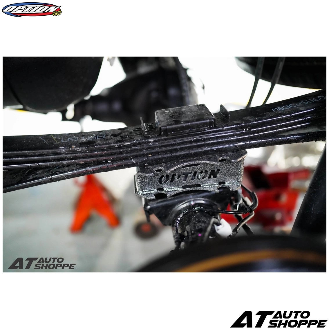 Close-up of a car suspension system with 'Option' branding, blurred background with 'AT Auto Shoppe' branding.